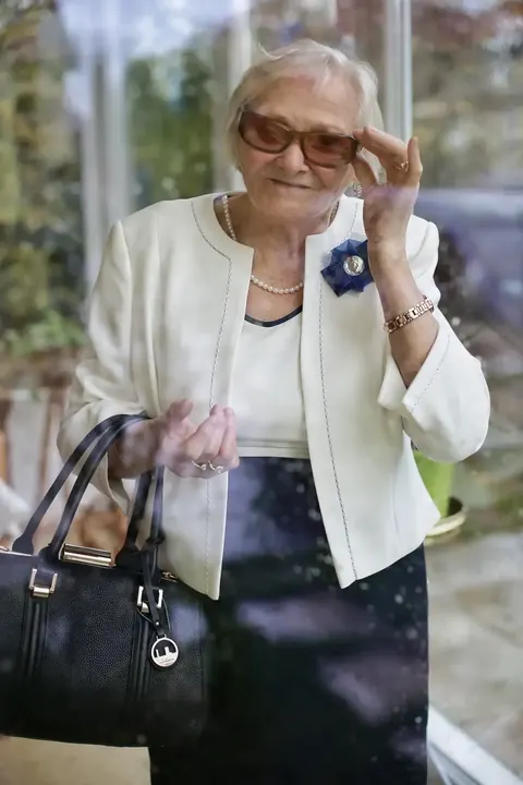 Umgebungsporträt einer elegant gekleideten älteren Frau mit schwarzer Handtasche im Freien; ihre gelöste Geste und das gepflegte Styling vermitteln Erfahrung…