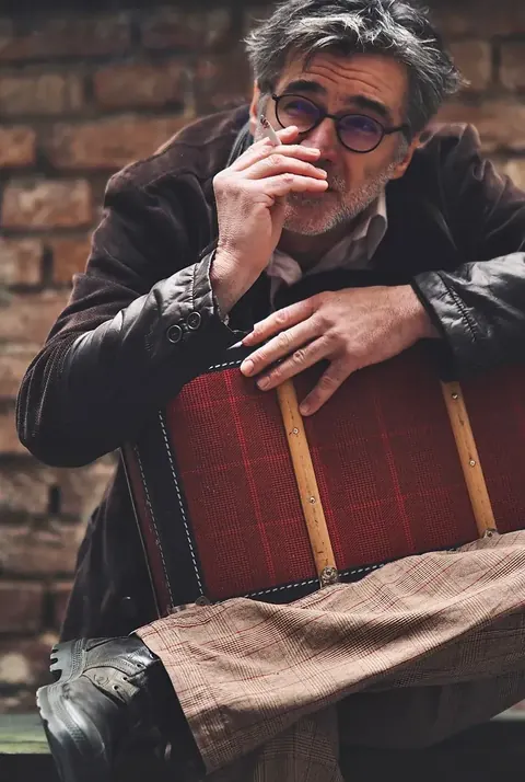 Charakterporträt eines sitzenden älteren Mannes mit Sonnenbrille, Lederjacke und Vintage-Koffer vor einer Ziegelwand; Texturen und Haltung formen eine filmische…