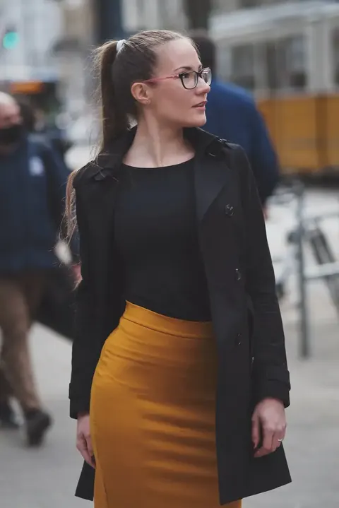 Urbanes Personal-Branding-Porträt einer Frau mit Brille, schwarzem Blazer und senffarbenem Rock, die über eine Straße blickt; die Komposition verbindet professionelle…
