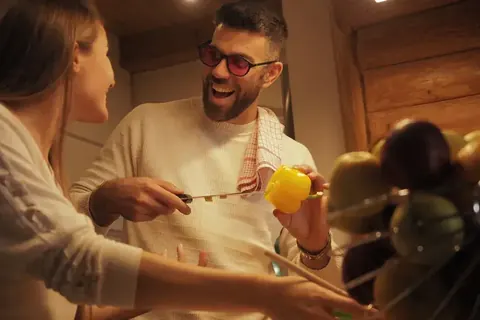 Warme Lifestyle-Szene eines lächelnden Mannes und einer Frau, die in einem hölzernen Interieur gemeinsam kochen; natürliche Gesten, Essenszubereitung und…