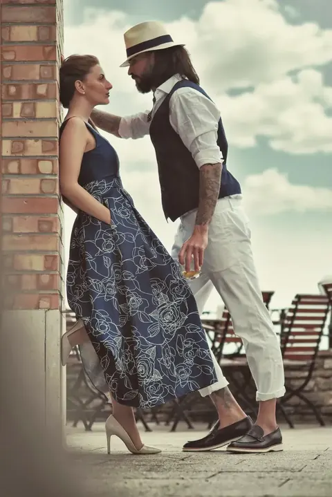Stilisiertes Paarporträt an einer Ziegelwand mit vintage-inspirierter Kleidung und enger Körpersprache; die Szene verbindet modisches Storytelling, Romantik und das…