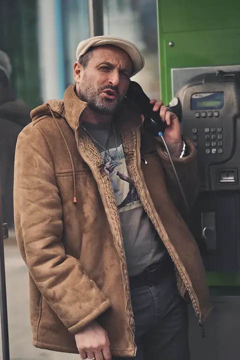 Straßen-Charakterporträt eines bärtigen Mannes mit Kappe und mehrlagiger brauner Kleidung an einem öffentlichen Telefon; gedämpfte Töne und verwitterte Texturen schaffen…
