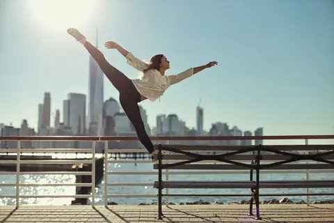 Tanzporträt auf einer Uferpromenade mit städtischer Skyline in der Ferne; die gestreckte Pose, geöffneten Arme und der tiefe Horizont drücken Disziplin, Freiheit und…