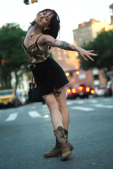 Dynamisches urbanes Fashionporträt einer Frau, die in der Dämmerung auf der Straße tanzt; Bewegung, Stadtlichter und entspanntes Styling erzeugen eine junge, unabhängige…