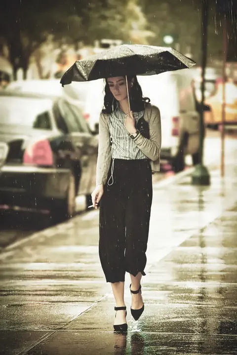 Editoriales Porträt einer Frau an einem Regentag unter einem dunklen Schirm; Spiegelungen, formelle Kleidung und gemessenes Tempo vermitteln Widerstandskraft und stille…