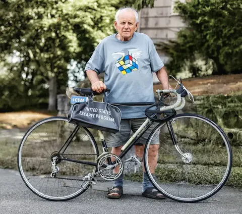 Dokumentarisches Porträt eines älteren Mannes mit Rennrad in einer Gartenumgebung; praktische Kleidung und direkte Darstellung rücken persönliche Geschichte und aktive…
