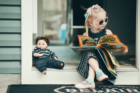 Erzählendes Porträt eines kleinen Kindes, das neben einer Puppe sitzt und in einem Türrahmen ein farbiges Buch liest; Styling und Requisiten schaffen eine spielerische…