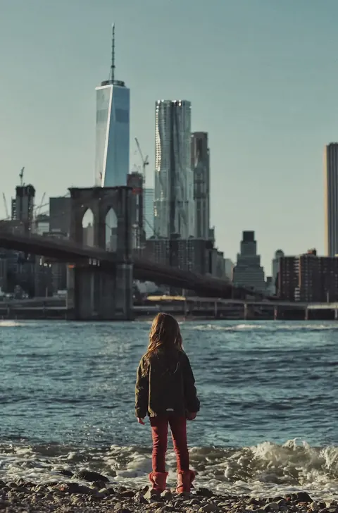 Kleine Figur mit Blick über das Wasser auf die Skyline von Manhattan; der monumentale Maßstab der Stadt macht die Szene zu einer visuellen Aussage über Ehrgeiz, Distanz…