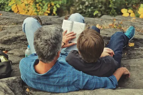 Familienbild aus der Vogelperspektive mit einem Erwachsenen und einem Kind, die im Freien liegend gemeinsam lesen; die enge Rahmung und die herbstliche Umgebung betonen…