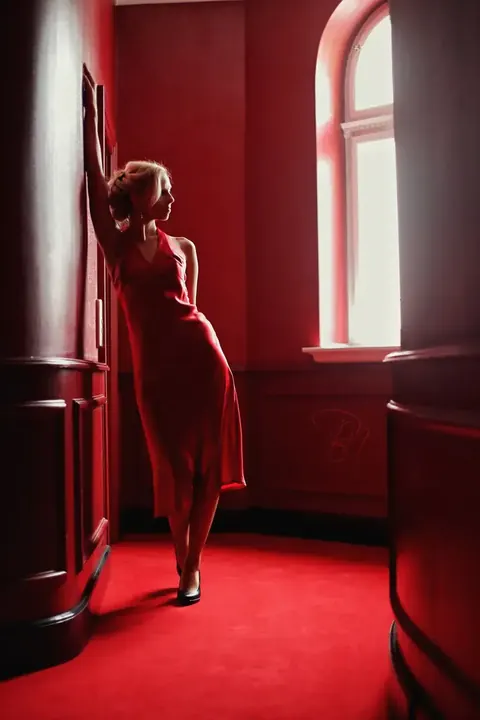 Dramatisches Fashionporträt einer Frau im roten Kleid zwischen hohen Fenstern in einem roten Interieur; Farbwiederholung und vertikale Architektur erzeugen theatralische…