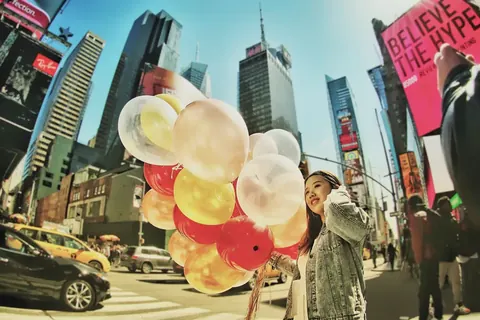 Lifestyle-Porträt am Times Square einer Frau mit einem Bündel transparenter Luftballons; leuchtende Stadtreklame und schwebende Formen schaffen Optimismus, Spektakel und…