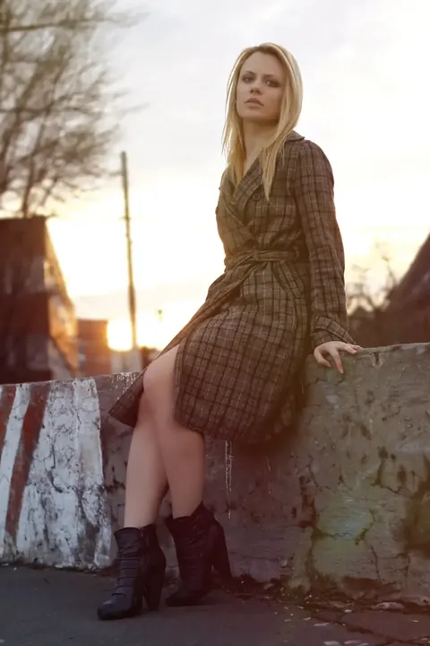 Umgebungs-Fashionporträt einer Frau auf einer Steinmauer bei Sonnenuntergang; gemustertes Kleid, Stiefel und tiefes warmes Licht vermitteln nachdenkliche Unabhängigkeit…