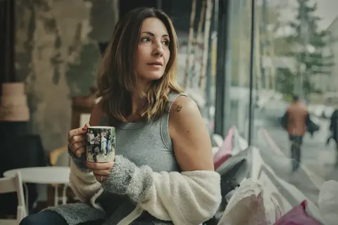 Natürliches Innenraumporträt einer Frau mit gemusterter Tasse am Fenster; mehrlagige Kleidung, weiches Tageslicht und aufmerksamer Blick vermitteln Wärme, Intelligenz und…