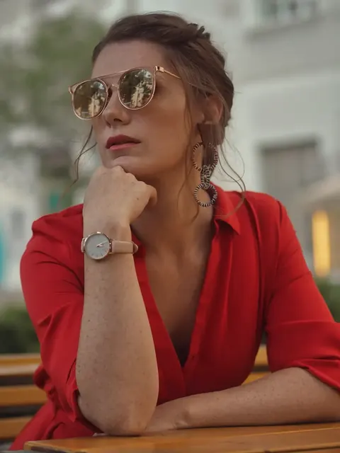 Nahes Editorialporträt einer Frau im roten Kleid und mit Sonnenbrille, das Kinn auf die Hand gestützt; die zurückhaltende Geste und die Accessoires vermitteln gefasste…