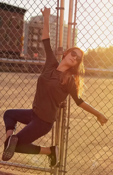 Urbanes Fashionporträt einer Frau, die sich im späten Sonnenlicht an einen Maschendrahtzaun lehnt; schräge Haltung und industrielle Textur schaffen eine rebellische…