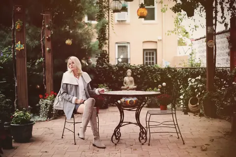 Editorialporträt einer Frau an einem Gartentisch in einem blumenreichen Innenhof; sanfte Farben und gelöste Haltung schaffen eine elegante, private und kultivierte…