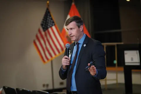 Keynote-Speaker mit Handmikrofon vor US-amerikanischen und österreichischen Flaggen; das Bild verbindet diplomatischen Kontext, Autorität und aktive Kommunikation.