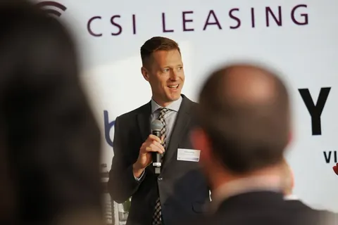 Corporate-Speaker über die Schulter eines Publikumsmitglieds bei einer gebrandeten Leasingveranstaltung fotografiert; die gestaffelte Tiefe bewahrt zugleich die Autorität…
