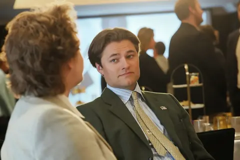 Ungezwungenes Gespräch bei einem Wirtschaftsempfang zwischen einem sitzenden Mann im Anzug und einer von hinten gesehenen Frau; konzentrierte Mimik und umstehende Gäste…