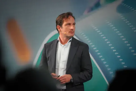 Konferenzporträt eines männlichen Vortragenden auf einer blau-grünen Bühne; kontrolliertes Licht und offene Haltung betonen Klarheit, Glaubwürdigkeit und professionelle…
