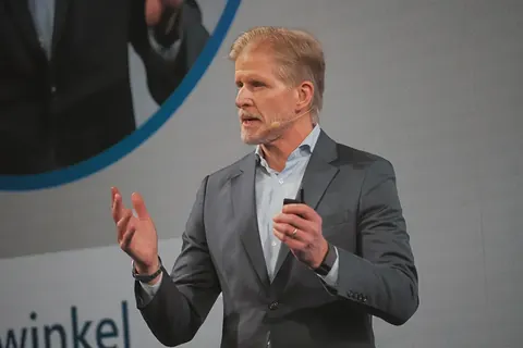 Nahaufnahme eines erfahrenen Redners auf der Bühne, der mit Mikrofon in der Hand gestikuliert; das Bild konzentriert sich auf Ausdruck, Argumentation und…