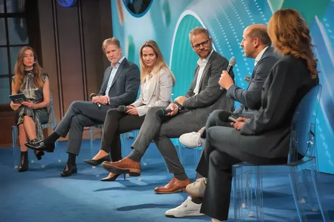 Podiumsdiskussion auf einer blauen Bühne mit sechs Teilnehmenden und Moderation; sitzende Interaktion, Mikrofone und publikumsoffene Komposition dokumentieren ein…