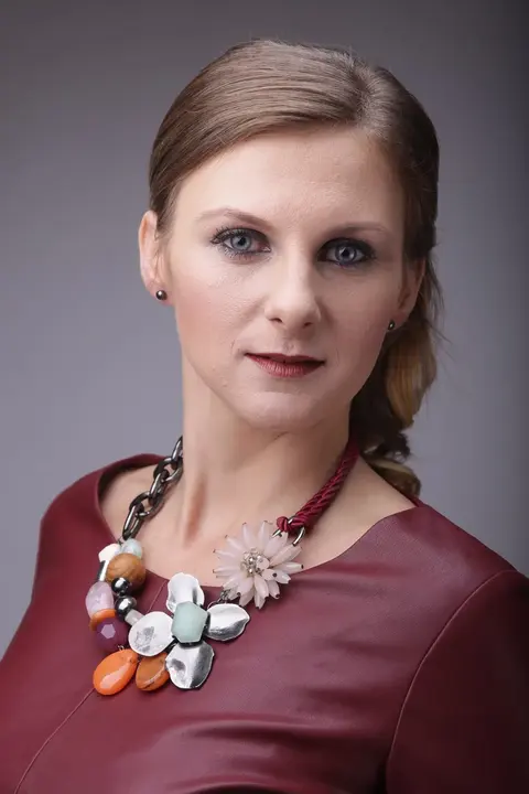 Formelles Studio-Headshot einer Frau mit sorgfältig frisiertem Haar, burgunderfarbener Kleidung und markanter Halskette; neutrales Licht lenkt die Aufmerksamkeit auf…