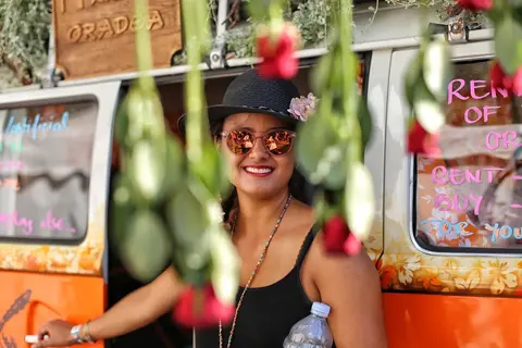 Farbiges Umgebungsporträt einer lächelnden Frau an einem städtischen Markt- oder Essensstand, teilweise von hängenden Blumen gerahmt; die Szene vermittelt Offenheit und…