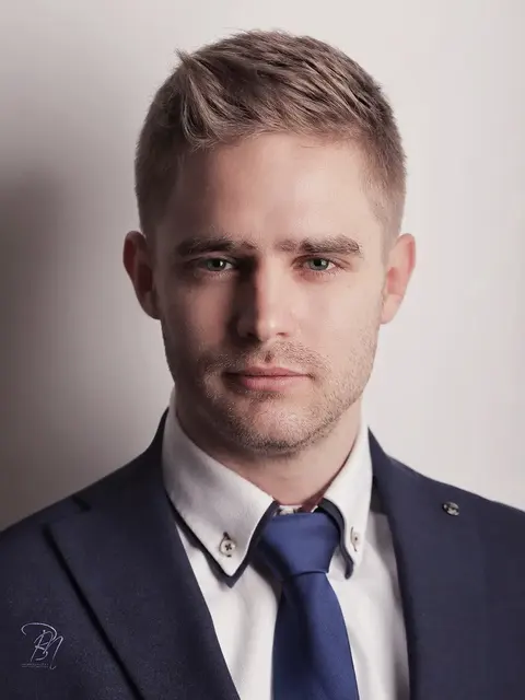 Klares Executive-Headshot eines jungen Mannes im dunklen Anzug und blauer Krawatte vor neutralem Hintergrund; ausgewogenes Licht und direkter Blick vermitteln…