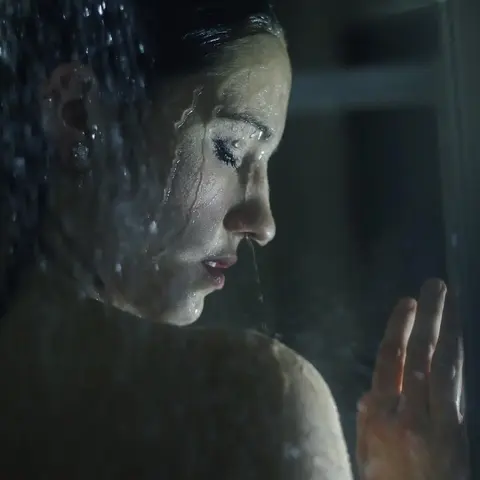 Stimmungsvolles Profilporträt einer Frau durch regennasses Glas; kühle Farben, Spiegelung und erhobene Hand erzeugen filmische Distanz und Introvertiertheit.