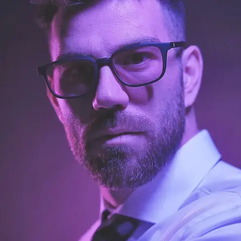 Low-Key-Porträt eines bärtigen Mannes mit Brille und formeller Kleidung unter violettem Licht; Farbkontrast und enger Ausschnitt schaffen eine zeitgenössische Executive-…