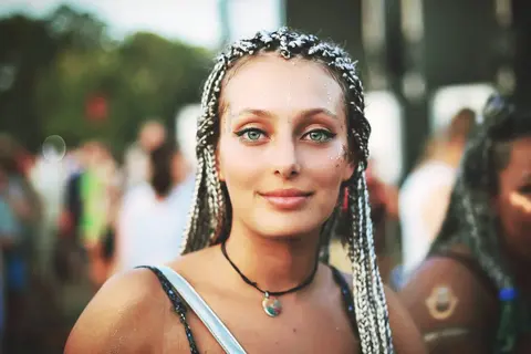 Straßenporträt einer Frau mit geflochtenem Haar und kräftigem Augen-Make-up bei einer öffentlichen Veranstaltung; geringe Schärfentiefe isoliert einen selbstbewussten…