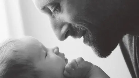 Zarte Schwarz-Weiß-Nahaufnahme eines erwachsenen Mannes, der sein Gesicht an die Stirn eines Babys legt; minimale Rahmung konzentriert sich auf Vertrauen, Schutz und…
