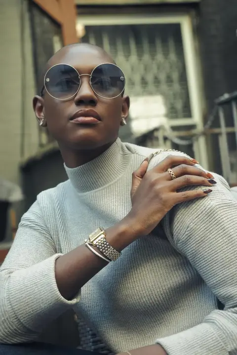 Urbanes Fashionporträt einer Frau mit runder Sonnenbrille und hellem Strickoberteil, die Hände nahe der Schulter; tiefe Perspektive und architektonischer Hintergrund…