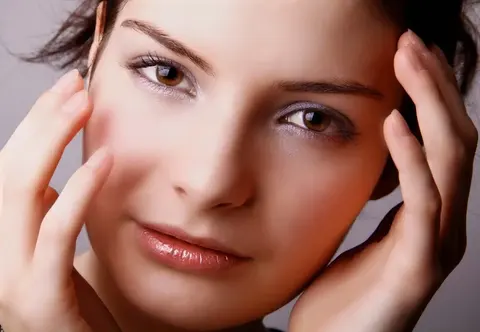 Beauty-Porträt eines Frauengesichts, das von beiden Händen vor klarem Hintergrund gerahmt wird; gleichmäßiges Licht, direkter Blick und sorgfältiges Styling rücken…