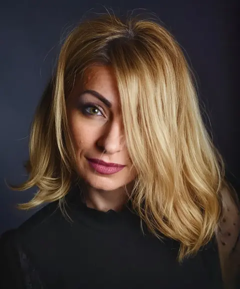 Low-Key-Studioporträt einer blonden Frau, deren Haar über ein Auge fällt; gerichtetes Licht und dunkler Hintergrund erzeugen Selbstbewusstsein, Geheimnis und…