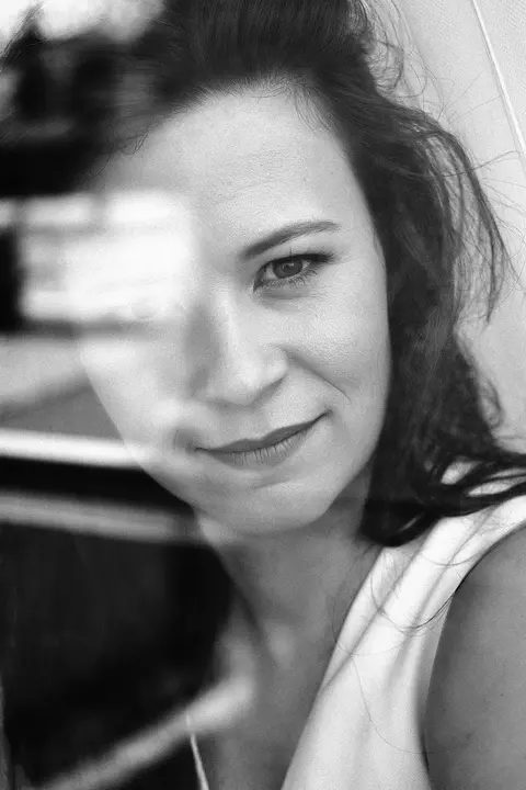 Geschichtetes Porträt einer Frau durch Glas oder Spiegelung mit einem scharf sichtbaren Auge; der fragmentierte Vordergrund deutet Beobachtung, Privatsphäre und komplexe…