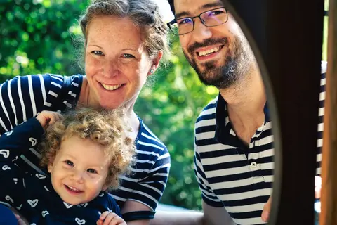 Helles Familienporträt zweier Erwachsener und eines lächelnden kleinen Kindes im Freien; abgestimmte gestreifte Kleidung und enge Gruppierung vermitteln Wärme…