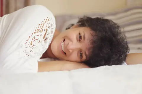 High-Key-Lifestyle-Porträt einer lächelnden Frau mit kurzem lockigem Haar auf einem Bett; weiße Texturen und gelöste Haltung vermitteln Komfort, Offenheit und Freude.