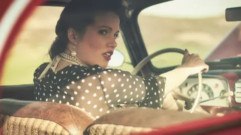 Vintage-inspiriertes Porträt einer Frau im gepunkteten Kleid und mit Handschuhen am Steuer eines klassischen Autos; Styling und warme Farben formen eine Erzählung von…