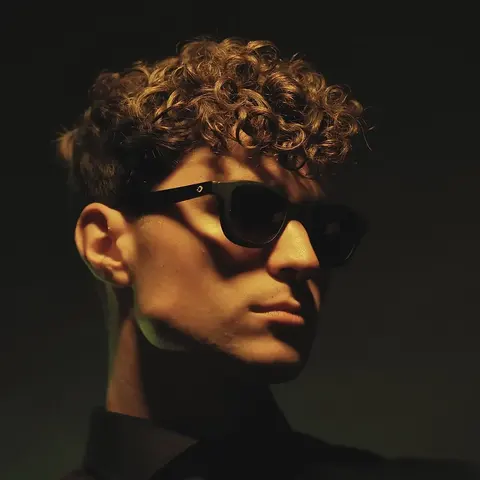 Low-Key-Studioporträt eines jungen Mannes mit lockigem Haar und Sonnenbrille im Seitenlicht; modellierter Schatten und minimales Styling schaffen eine starke Musik- oder…