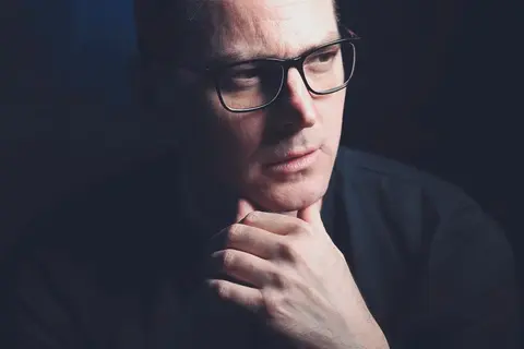 Nahes Executive-Porträt eines Mannes mit Brille und der Hand am Kinn; dunkler Hintergrund und nachdenklich gesenkter Blick vermitteln Analyse, Erfahrung und Diskretion.