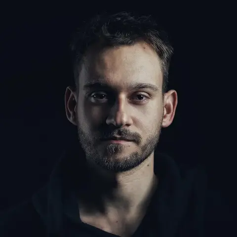 Low-Key-Headshot eines Mannes vor Schwarz, wobei eine Gesichtshälfte aus dem Schatten tritt; direkter Blick und minimales Styling vermitteln Fokus, Ernsthaftigkeit und…