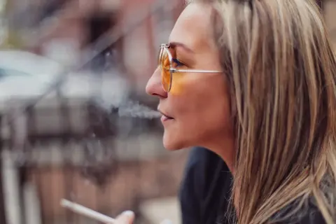 Ungezwungenes Seitenprofil einer Frau mit bernsteinfarbener Brille in urbaner Umgebung; geringe Schärfentiefe und natürliche Haltung vermitteln Aufmerksamkeit…